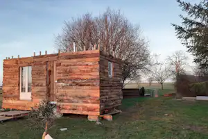 Holzhütte auf einem Campingplatz mit Fenster und Tür, umgeben von Bäumen und Wiese im Hintergrund.