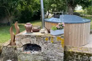 Holz-Whirlpool im Garten mit Steinmauer, Blumen im Kasten und Feuerstelle, umgeben von Bäumen und Rasen.