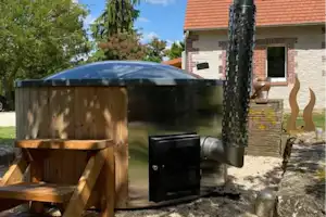 Holzfass-Sauna mit Kamin und Ofen vor einem Backsteingebäude, umgeben von Bäumen und Rasen.
