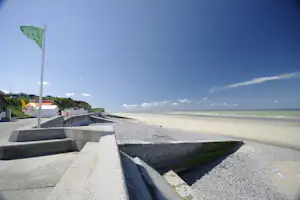 Sandstrand am Meer, Blick von einem Campingplatz mit Betonmauer, Kiesweg und Fahnenmast im Vordergrund.