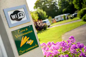 WLAN-Schild am Campingplatz an einer Hauswand, im Hintergrund Rasen, Wohnwagen und lila Blumenbeet.