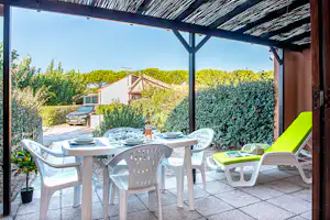 Terrasse mit weißer Eßtischgruppe, grünen Liegen und Hecke, im Hintergrund ein geparktes Auto und Wohnhaus.
