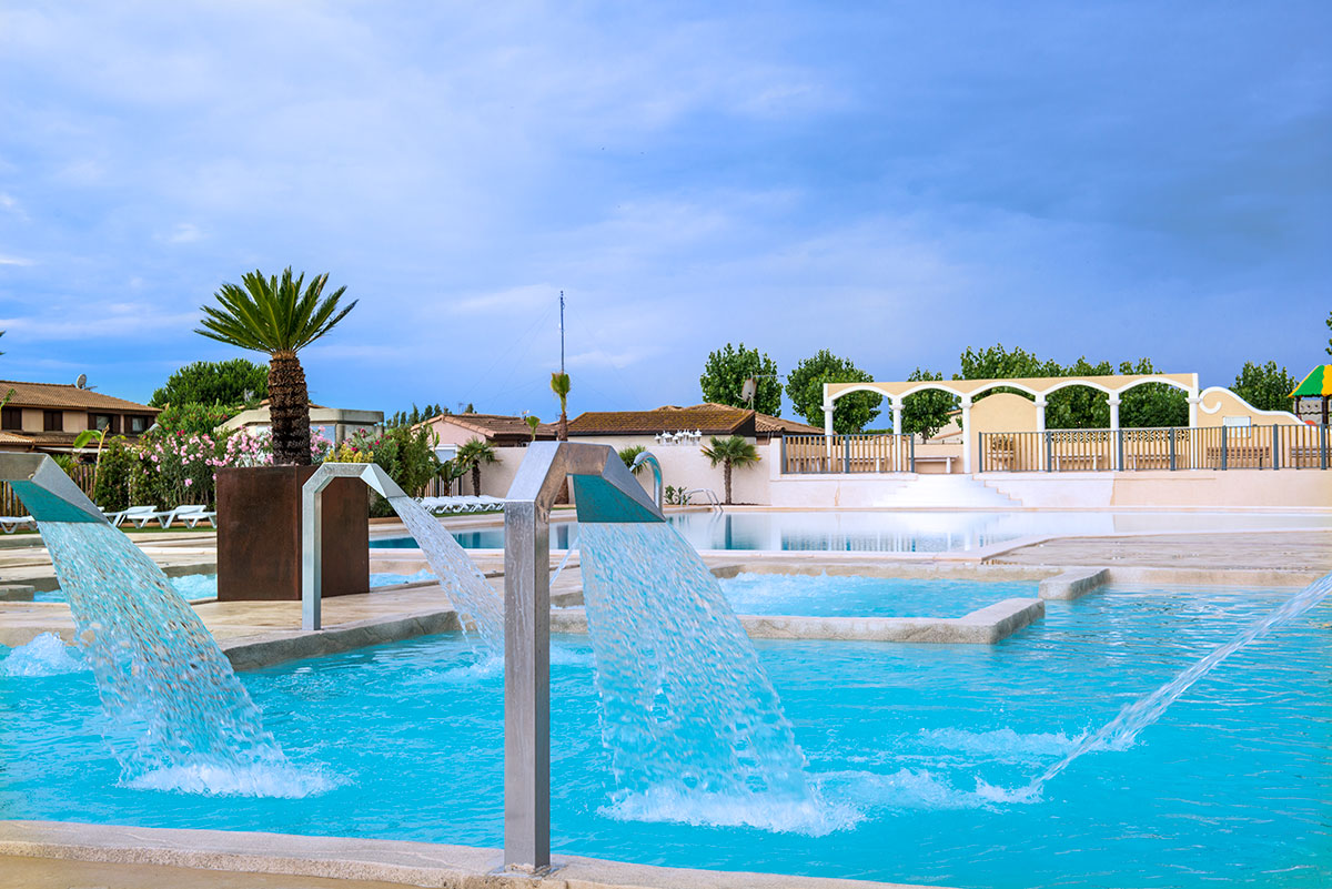 Beheizter Camping-Pool mit mehreren Liegen, weißen Umrandungen und Zaun, im Hintergrund Palmen und Gebäude.