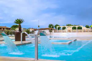 Beheizter Camping-Pool mit mehreren Liegen, weißen Umrandungen und Zaun, im Hintergrund Palmen und Gebäude.