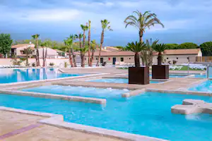 Großer Camping-Pool mit mehreren Becken, Palmen und weißen Gebäuden im Hintergrund, Liegeflächen und Wege am Rand.