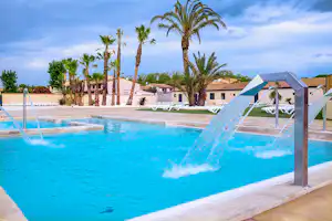 Blauer Camping-Pool mit Liegen am Beckenrand, Palmen und weißen Gebäuden im Hintergrund.