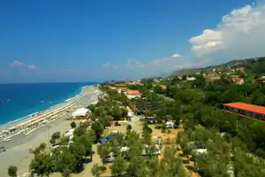 Blick auf einen Campingplatz an der Küste mit Meer, Strandpromenade, Bäumen und Häusern am Hang.