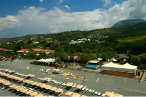 Blick auf einen Campingplatz mit Pool, Liegen am Strandbereich und Wohngebäuden, umgeben von grünen Hügeln und Häusern.
