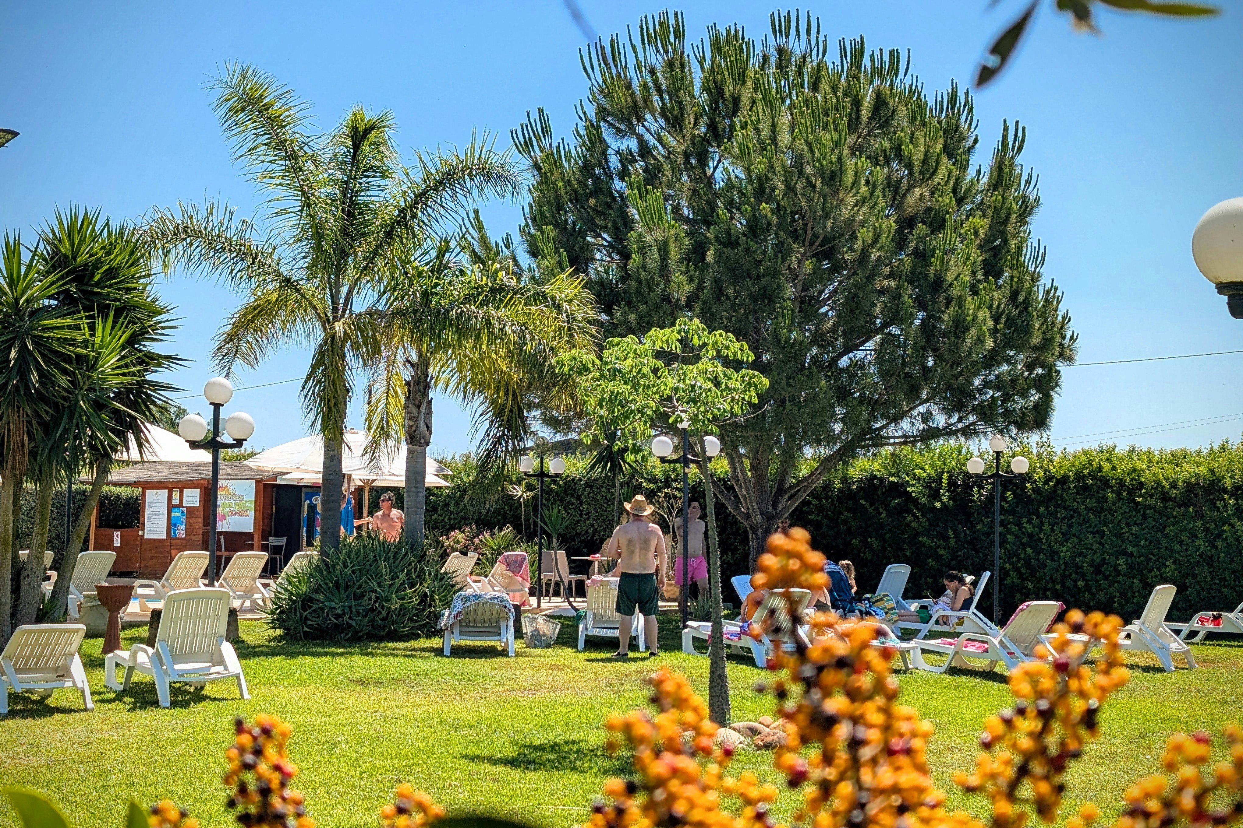 Gepflegter Campingplatz mit Rasen, Liegestühlen und Sonnenschirmen, im Hintergrund Bäume und ein Gebäude, im Vordergrund Blumen.