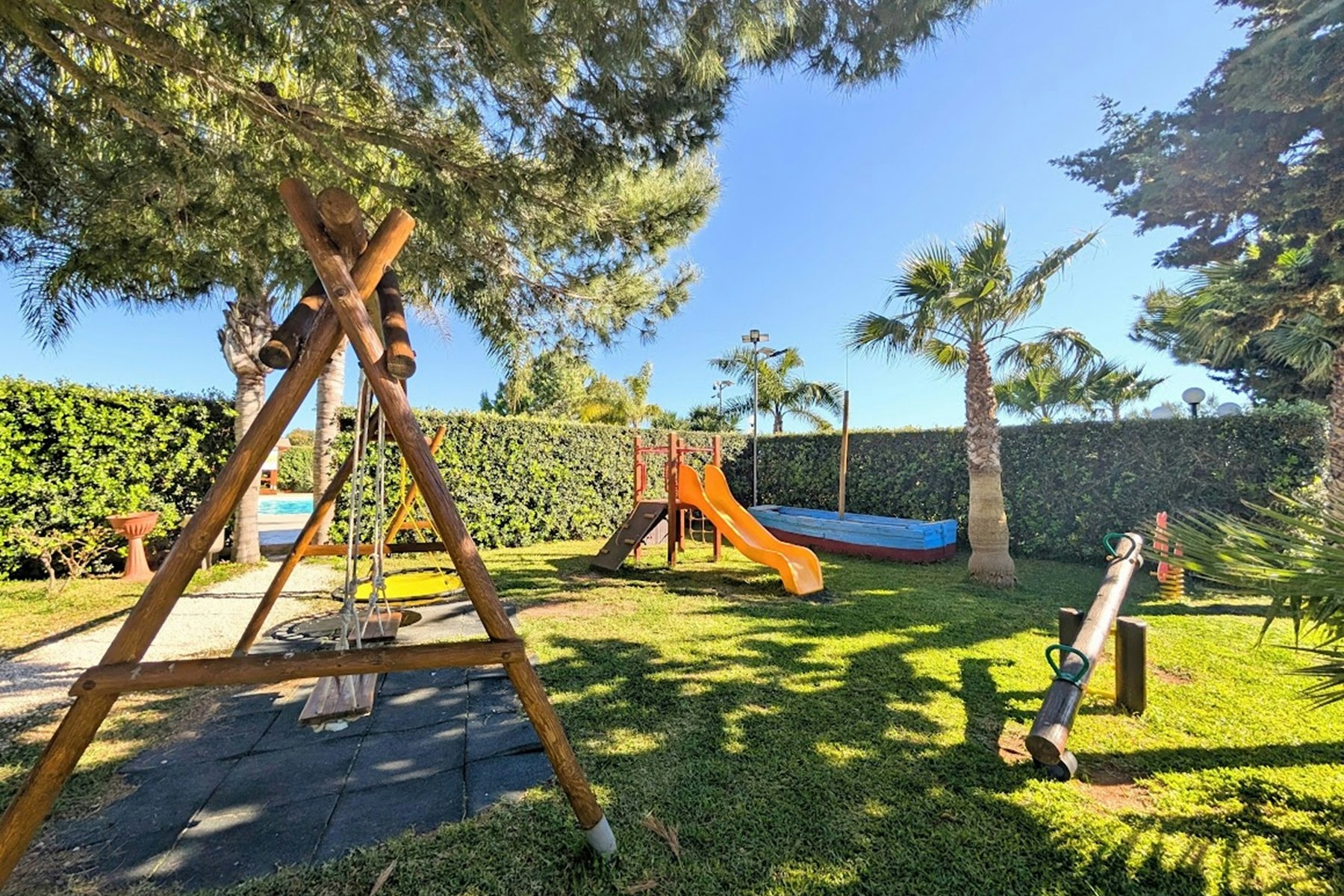 Spielplatz im Garten: Schaukelgestell mit Rutsche, Trampolin und Rasenfläche, umgeben von Hecke, Palmen und Bäumen.