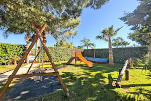 Spielplatz im Garten: Schaukelgestell mit Rutsche, Trampolin und Rasenfläche, umgeben von Hecke, Palmen und Bäumen.