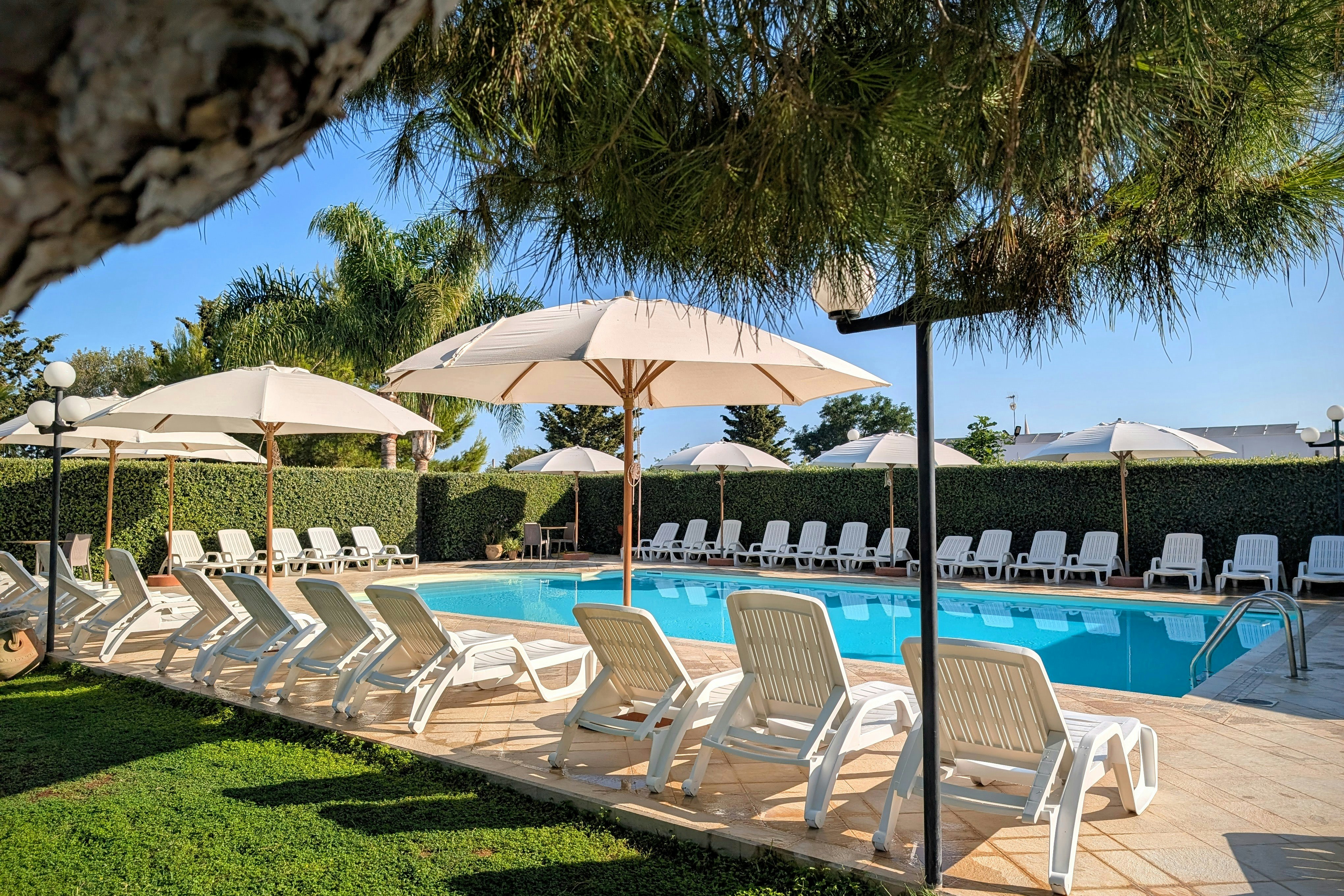 Reihe Sonnenliegen am Pool mit Sonnenschirmen, gepflegter Rasenfläche und Hecken, im Hintergrund ein Fels und Bäume.