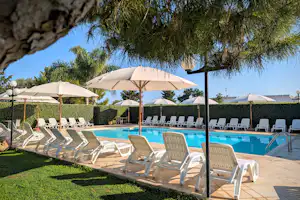 Reihe Sonnenliegen am Pool mit Sonnenschirmen, gepflegter Rasenfläche und Hecken, im Hintergrund ein Fels und Bäume.