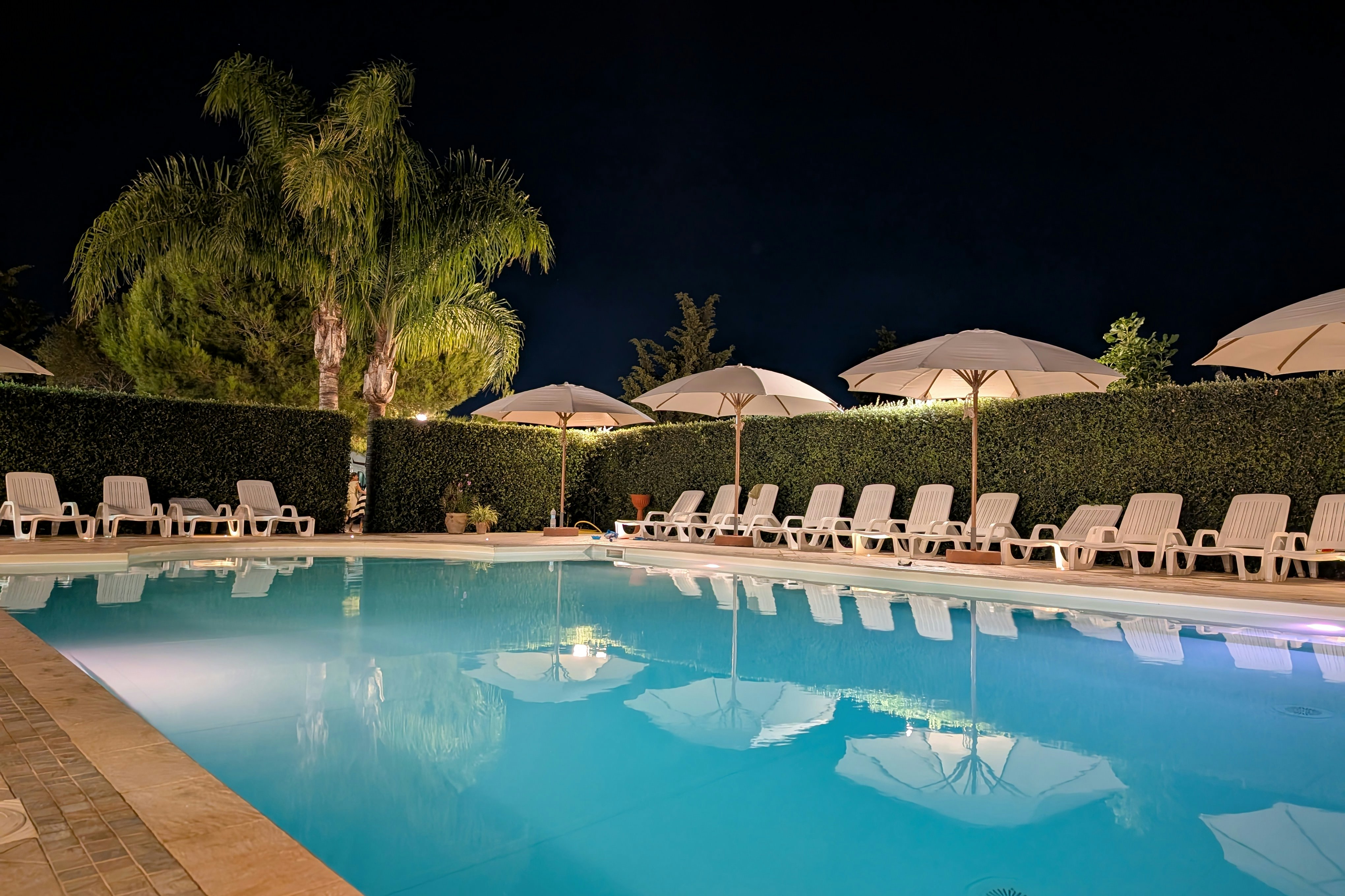 Schwimmbad mit Liegestühlen am Rand, dahinter Hecken und mehrere Sonnenschirme bei Nacht.