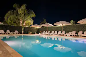 Schwimmbad mit Liegestühlen am Rand, dahinter Hecken und mehrere Sonnenschirme bei Nacht.