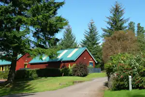 Rote Hütte mit grünem Dach auf einem Campingplatz, umgeben von Büschen, Rasen und hohen Nadelbäumen.