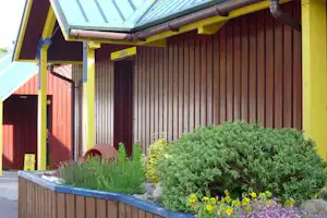Gelb gestrichene Ferienhaus-Fassade mit überdachtem Eingang, Holzverkleidung und gepflegtem Blumenbeet mit Sträuchern.