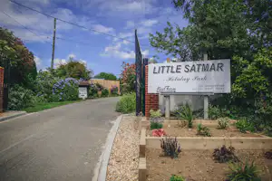 Einfahrt zum Campingplatz mit Schild „Little Satmar“, Ziegelpfeiler, Kiesbeet und Straße am Rand mit Bäumen und Sträuchern.
