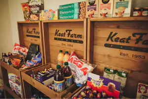 Regal mit Lebensmitteln und Getränken in einer kleinen Verkaufsbude, darunter Keat Farm Aufdrucke und Snacks in Tüten.