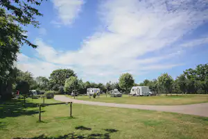 Campingplatz mit Wohnwagen auf Stellplätzen, breitem Weg und Rasenflächen, umgeben von Bäumen und Hecken.