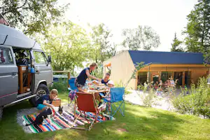 Wohnmobil am Campingplatz mit geöffnetem Fahrerhaus, Picknickdecke, Tisch und Stühlen vor einem Zelt und Grünfläche.