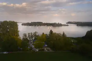 Blick auf einen See mit Uferpromenade, Campingplätzen und Wohnwagen zwischen Bäumen und grünen Hängen.