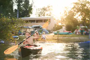 Ein Kajak auf dem Campingsee, im Hintergrund Zelte und weitere Boote, im Abendlicht zwischen Bäumen.