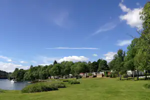Campingplatz am See mit Rasenfläche, Bäumen und Wohnwagen, im Hintergrund Häuser und Steg am Ufer.