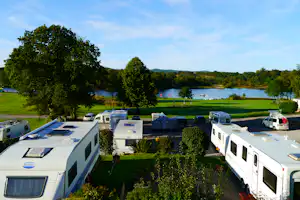 Wohnmobile und Autos auf dem Campingplatz am See, umgeben von Rasenflächen und Bäumen unter blauem Himmel.