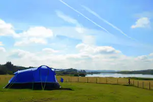 Blaues Campingzelt am Seeufer auf einer Wiese, im Hintergrund Felder, Bäume und ein weiter Himmel mit Wolken.