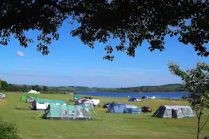 Campingplatz am See: Zelte und Wohnwagen auf einer Wiese, Autos und Wohnmobile, im Hintergrund Wasser und grüne Hügel.
