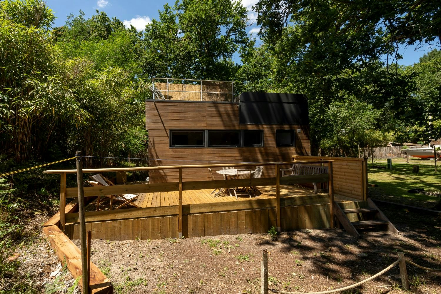Holz-Campingunterkunft mit Terrasse, Sitzbank und Stühlen, umgeben von Bäumen und hohem Gras im grünen Garten.