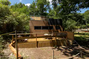 Holz-Campingunterkunft mit Terrasse, Sitzbank und Stühlen, umgeben von Bäumen und hohem Gras im grünen Garten.