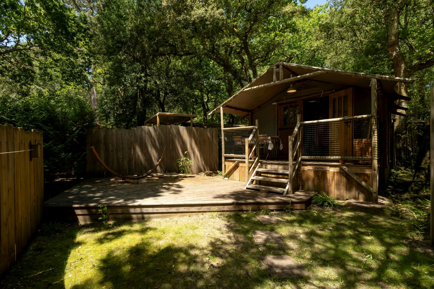 Holzterrasse mit Zeltunterkunft und Veranda, umgeben von hohen Bäumen und einem Holzsichtschutz am Campingplatz.