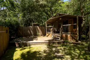 Holzterrasse mit Zeltunterkunft und Veranda, umgeben von hohen Bäumen und einem Holzsichtschutz am Campingplatz.