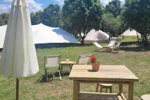 Weiße Sonnenschirm- und Holztisch-Sitzgruppe auf einer Wiese vor mehreren Zelten mit Liegen und Bäumen im Hintergrund.