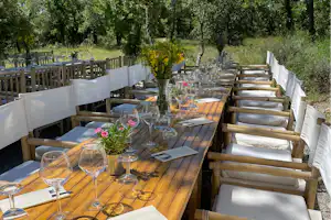 Festlich gedeckter Holztisch mit Gläsern und Blumen auf einer Terrasse, umgeben von weißen Stühlen und Bäumen im Grünen.