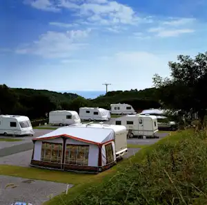 Zelt mit Glasfront und Wohnwagen auf einem Campingplatz, umgeben von Bäumen und grünen Hügeln im Hintergrund.