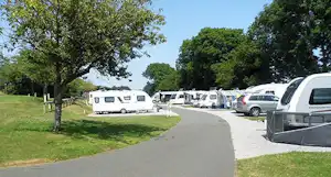 Wohnwagen und Autos auf einem Campingplatz, gepflasterter Weg zwischen Stellplätzen, Bäume und Rasen am Rand.