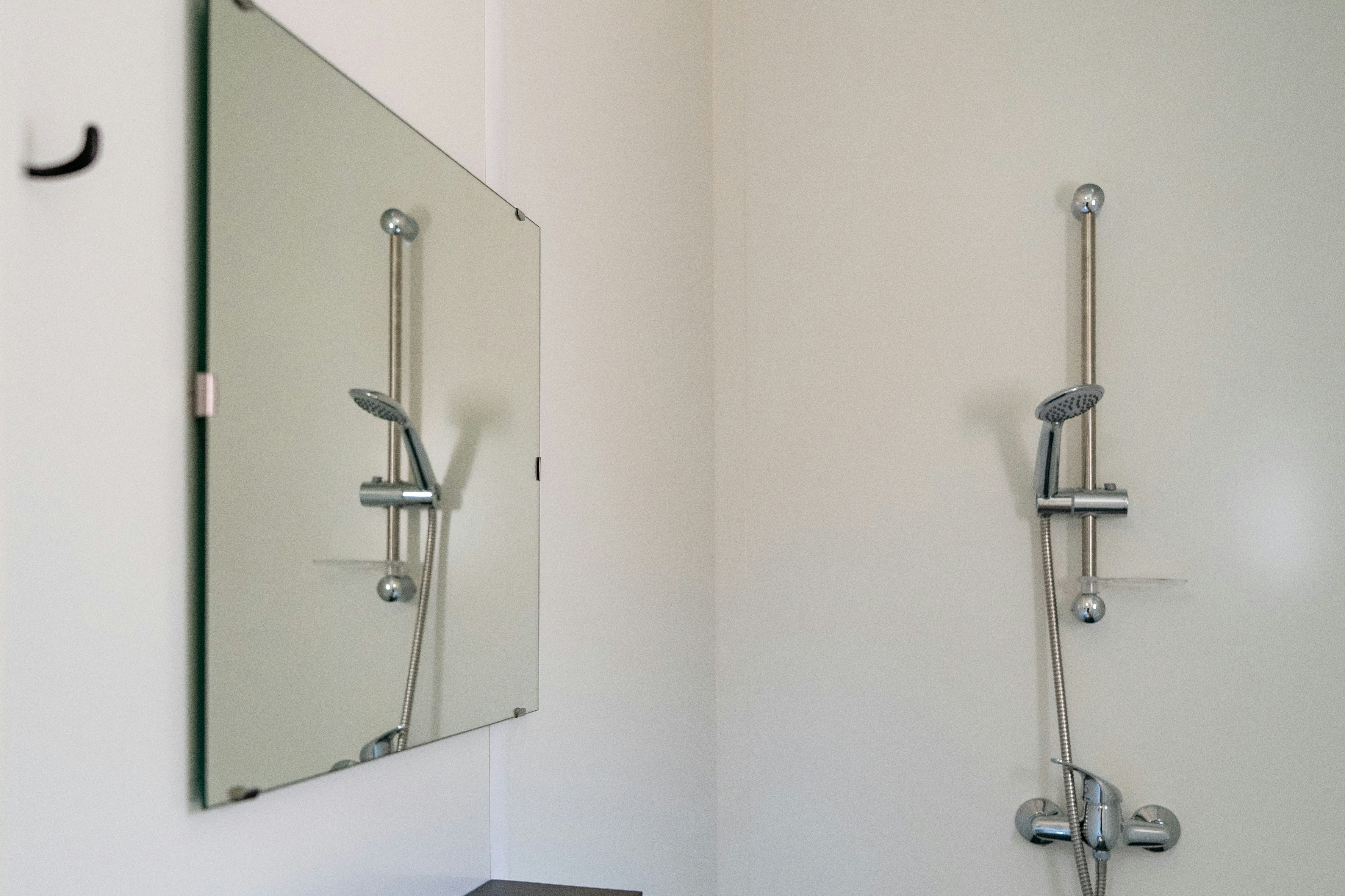 Modernes Badezimmer mit Waschbecken, Spiegel und Duscharmatur an der Wand, klare weiße Fliesen und Duschkopf.