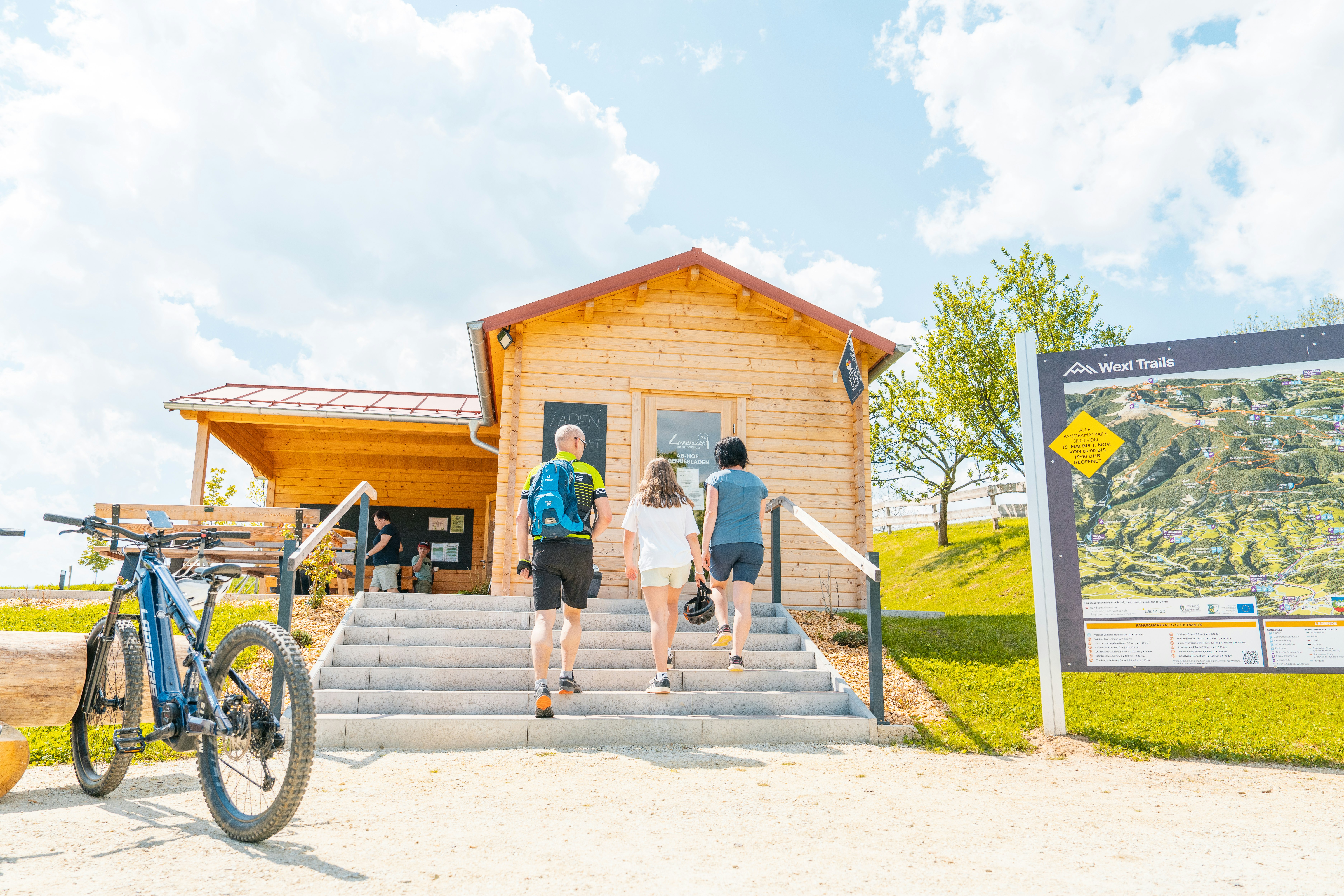 Holzgebäude mit Treppe und Fahrrädern davor, zwei Personen laufen am Eingang vorbei, daneben eine Infotafel auf dem Campingplatz.
