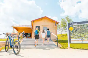 Holzgebäude mit Treppe und Fahrrädern davor, zwei Personen laufen am Eingang vorbei, daneben eine Infotafel auf dem Campingplatz.