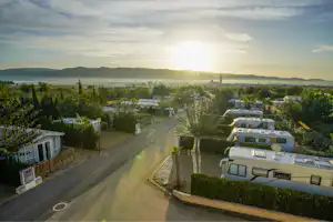 Campingplatz mit Wohnmobilen und Wohnwagen entlang eines Wegs, im Hintergrund Bäume, Hügel und die Sonne am Himmel.