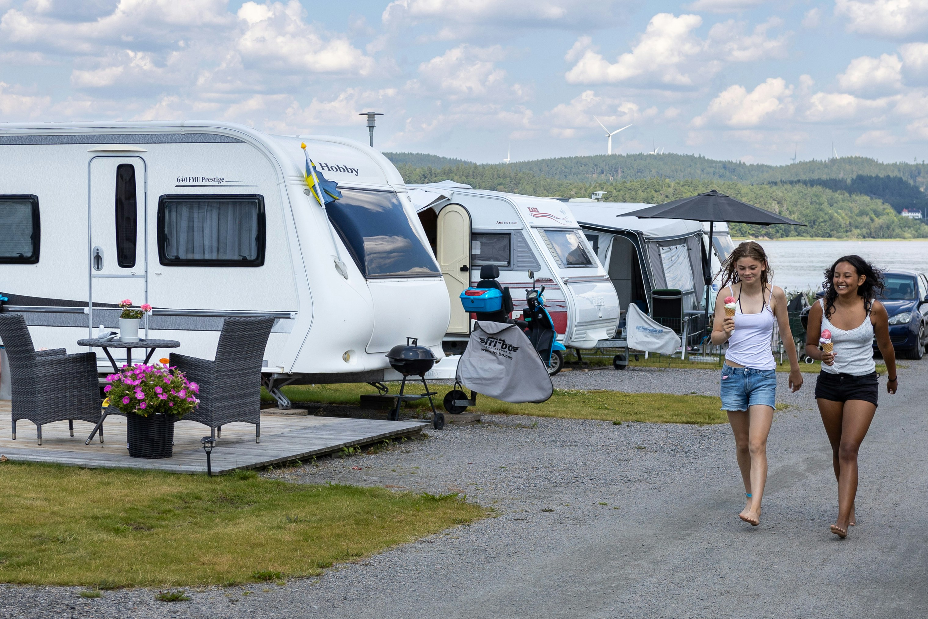 Wohnwagen und Campingfahrzeuge auf einem Stellplatz am See, mit zwei Personen auf dem Weg zwischen den Parzellen.