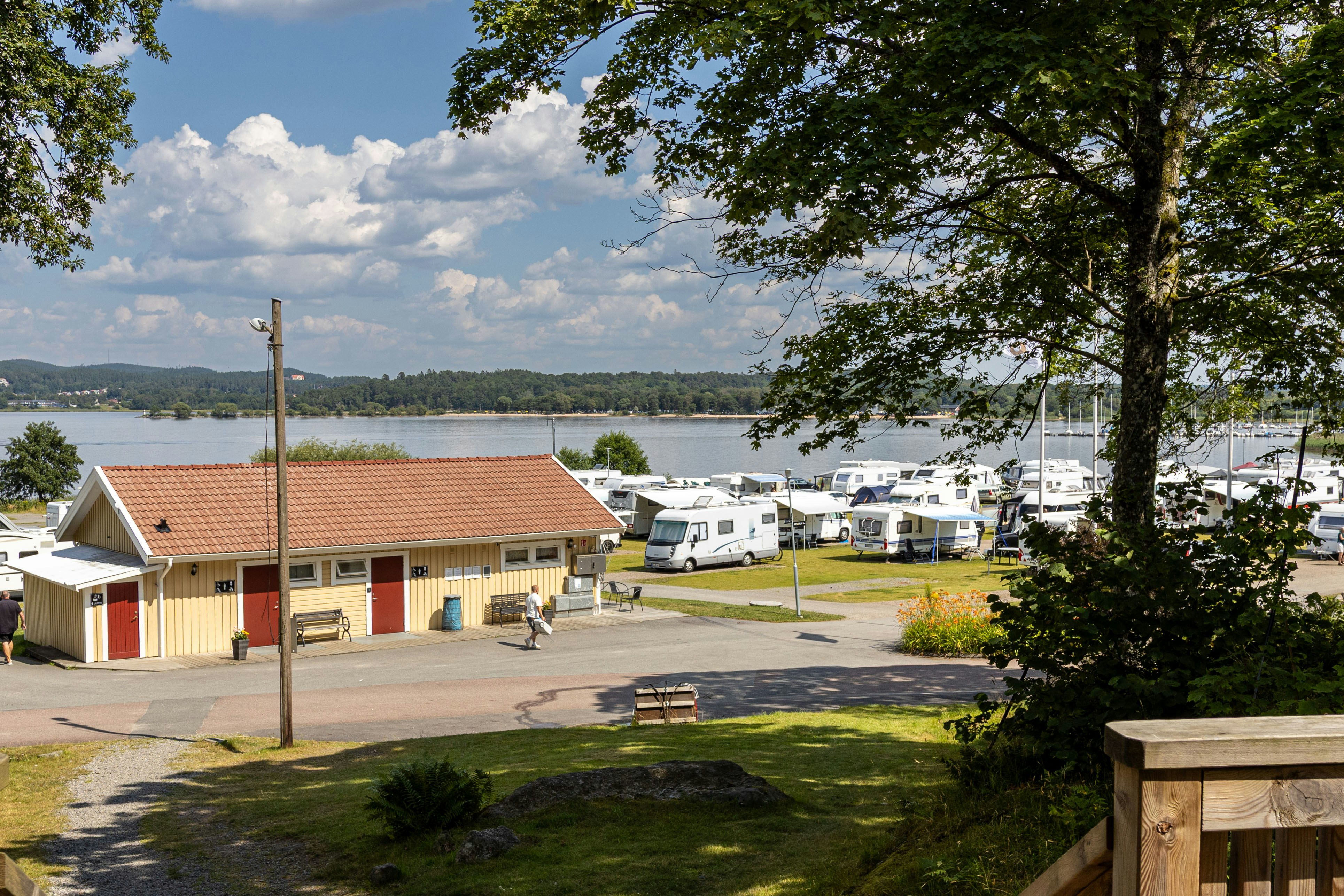 Campingplatz mit Wohnwagen und Mobilheimen, gepflasterter Straße, Bank und Bäumen am Rand, Blick auf den See in der Ferne.