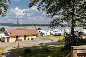 Campingplatz mit Wohnwagen und Mobilheimen, gepflasterter Straße, Bank und Bäumen am Rand, Blick auf den See in der Ferne.