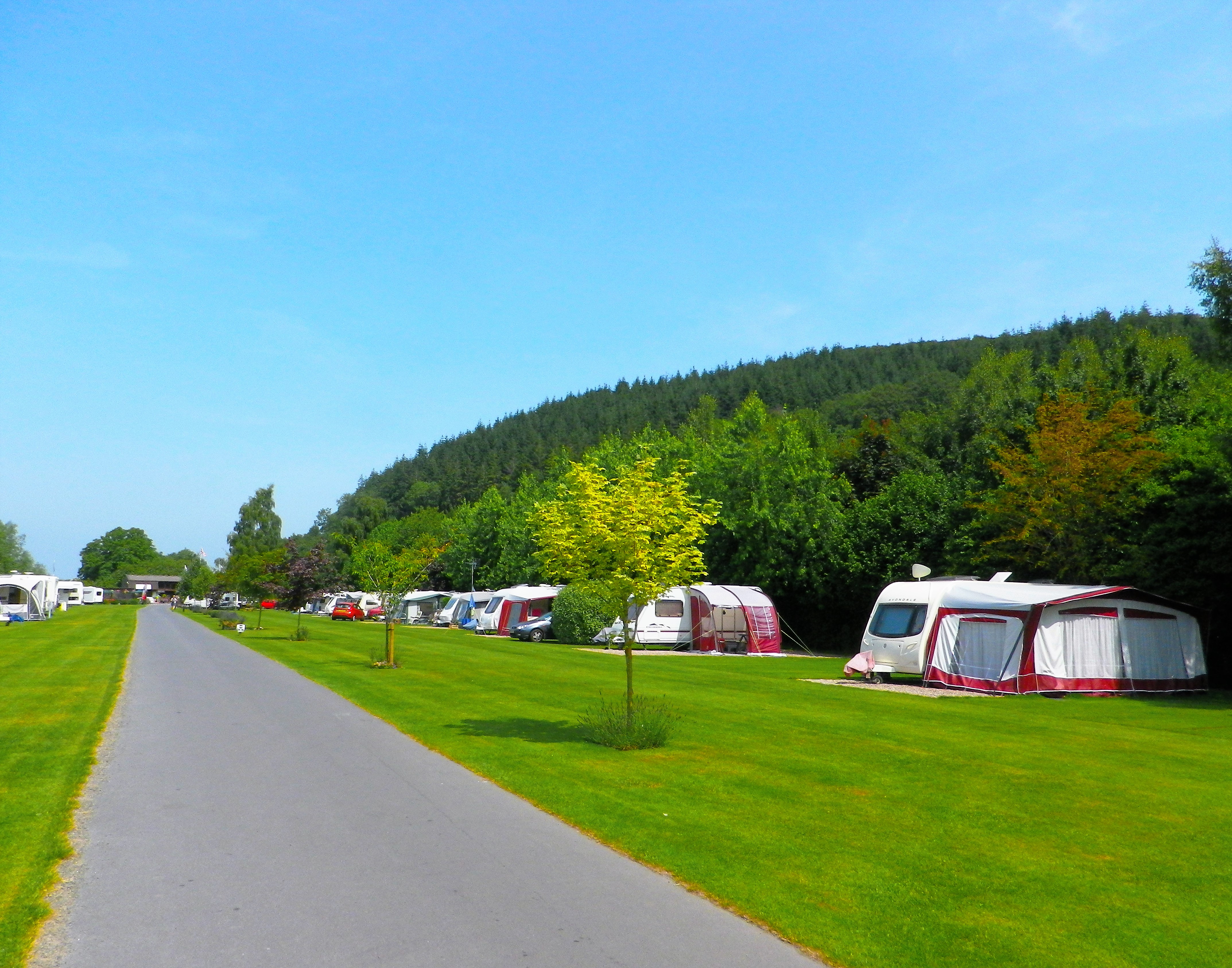 Reihe von Wohnwagen auf einem Campingplatz am grünen Rasen, mit breitem Weg und bewaldeten Hügeln im Hintergrund.