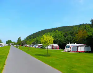 Reihe von Wohnwagen auf einem Campingplatz am grünen Rasen, mit breitem Weg und bewaldeten Hügeln im Hintergrund.