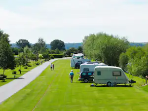 Wohnwagen und Campingbusse stehen auf einem grünen Stellplatz neben einem breiten Weg, umgeben von Bäumen und Rasen.