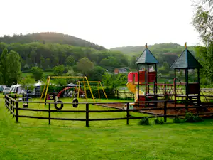 Spielplatz auf grünem Rasen mit Klettergerüst und Rutsche, im Hintergrund Bäume, Hügel und Wohnwagen am Campingplatz.